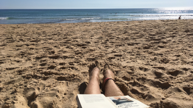 Person am Strand, man sieht nur ihre Beine. Sie hat ein Buch auf den Beinen liegen und schaut Richtung Meer